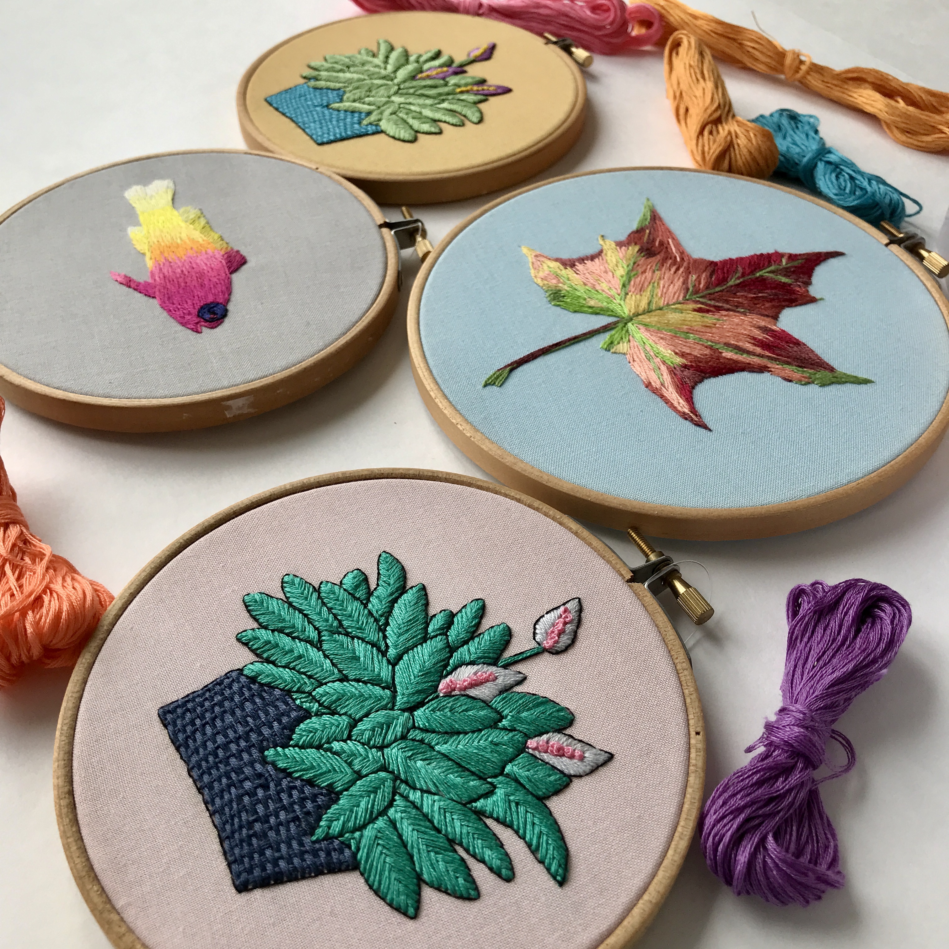 Four embroidery hoops with nature-inspired embroidery designs and colorful skeins of thread. Free stitch-alongs made  by @faimyxstitch