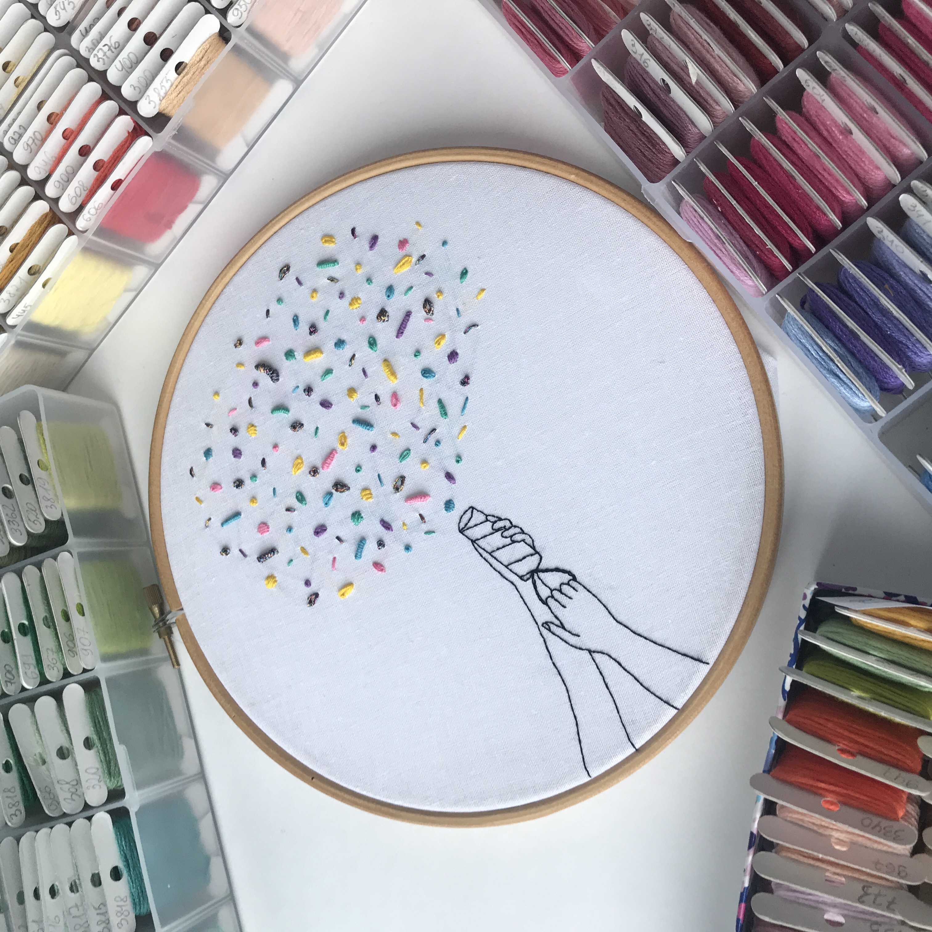 Embroidery hoop with line-drawn hands releasing a burst of colorful speckled stitches