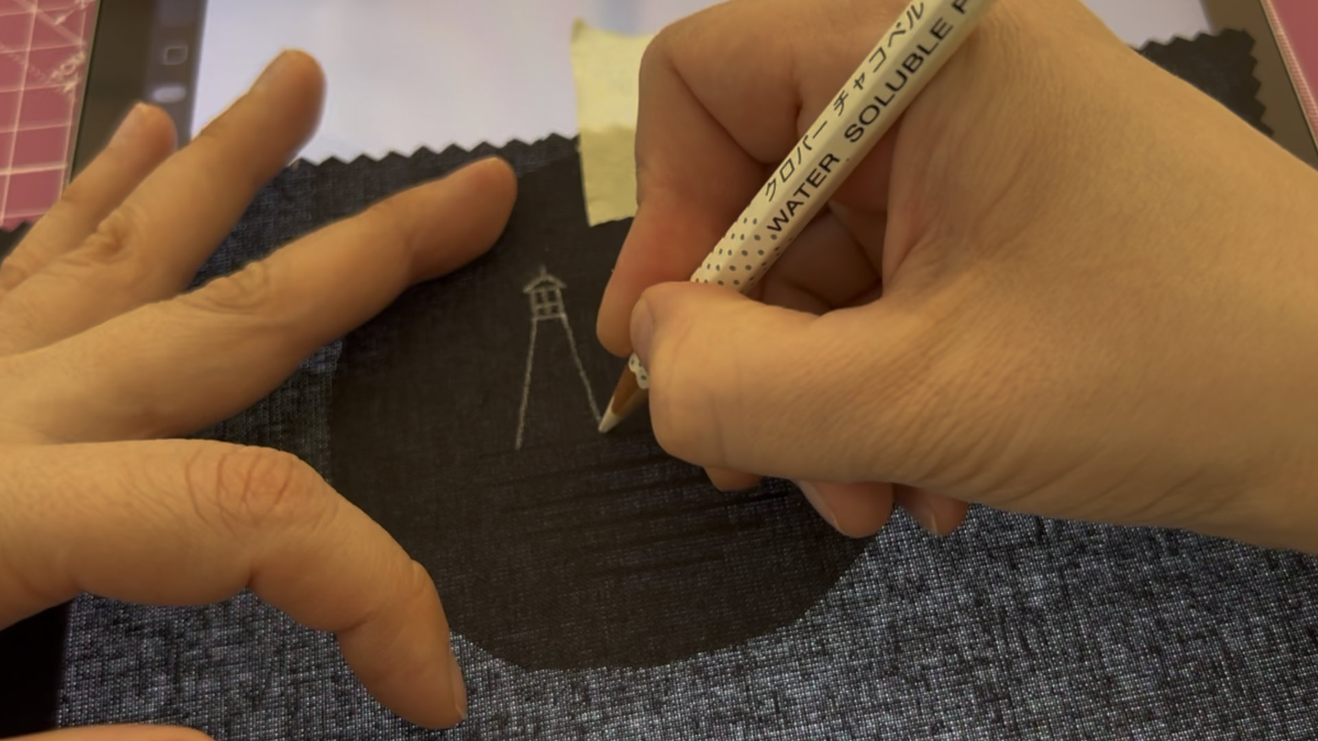 Close-up of hand sketching a tiny lighthouse on dark fabric with a water-soluble pencil.