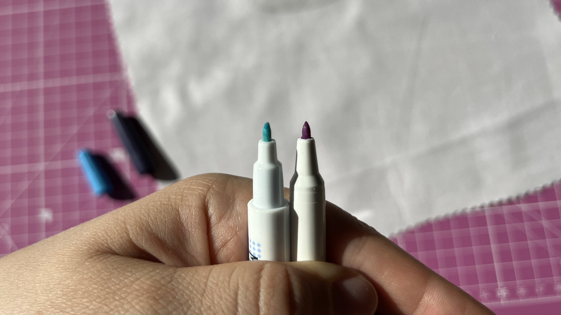 Two fabric marker tips (blue and purple) held by a hand, pink grid cutting-mat background.