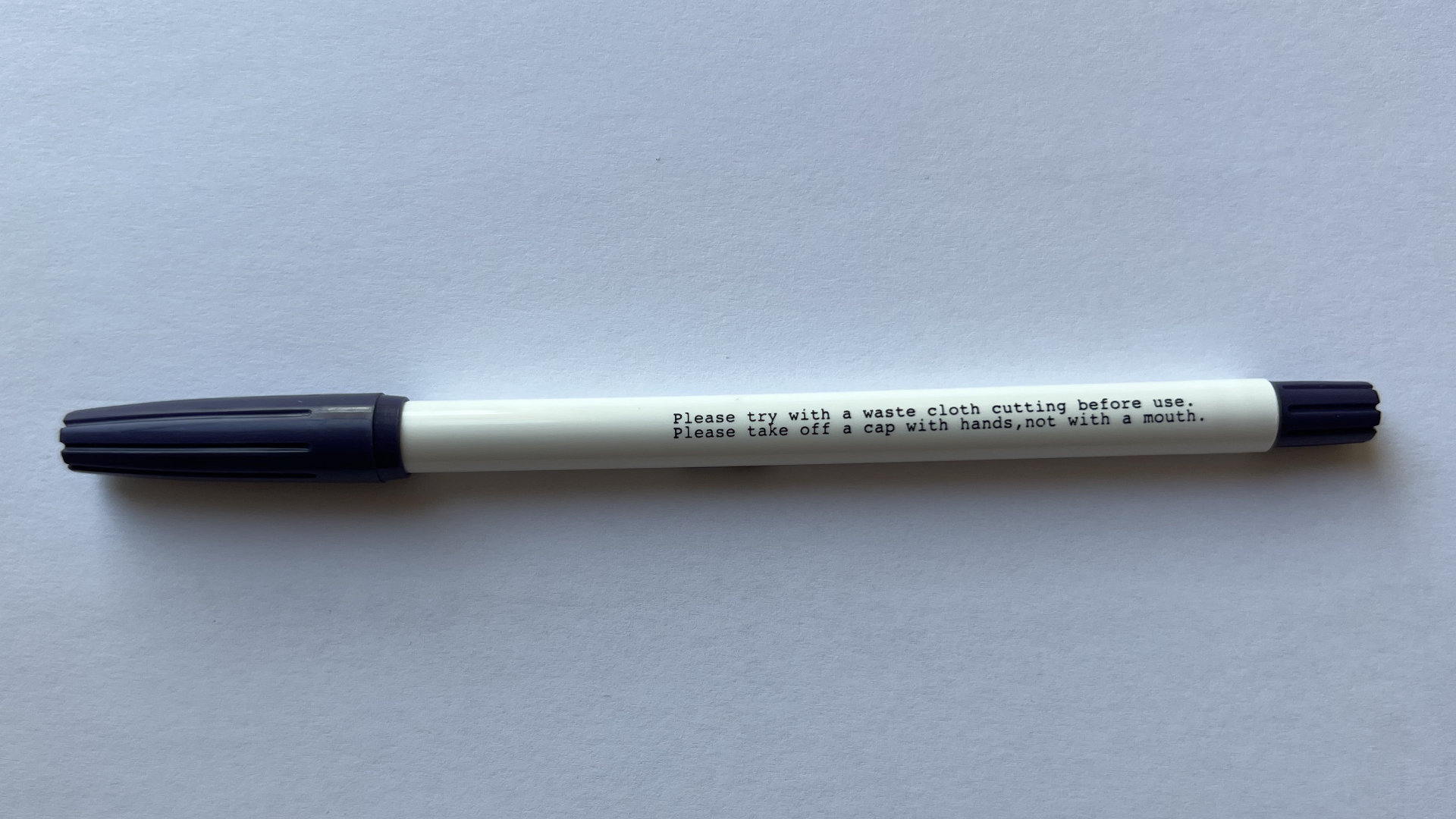 White marker pen with dark blue caps on both ends, resting on a light surface, with printed instructions along the body.