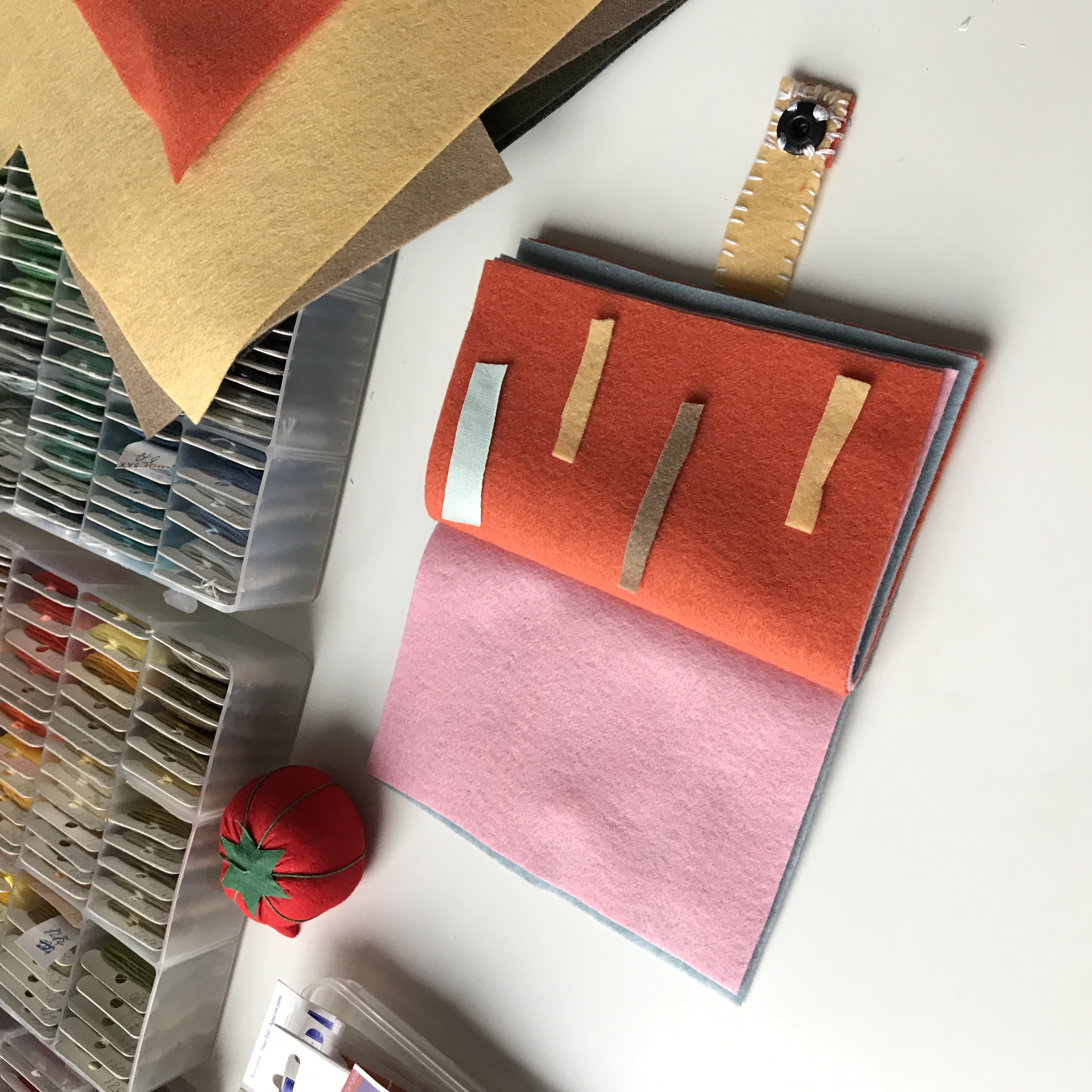 Open felt notebook with orange cover and stitched felt strips, pink inner page, craft supplies in background