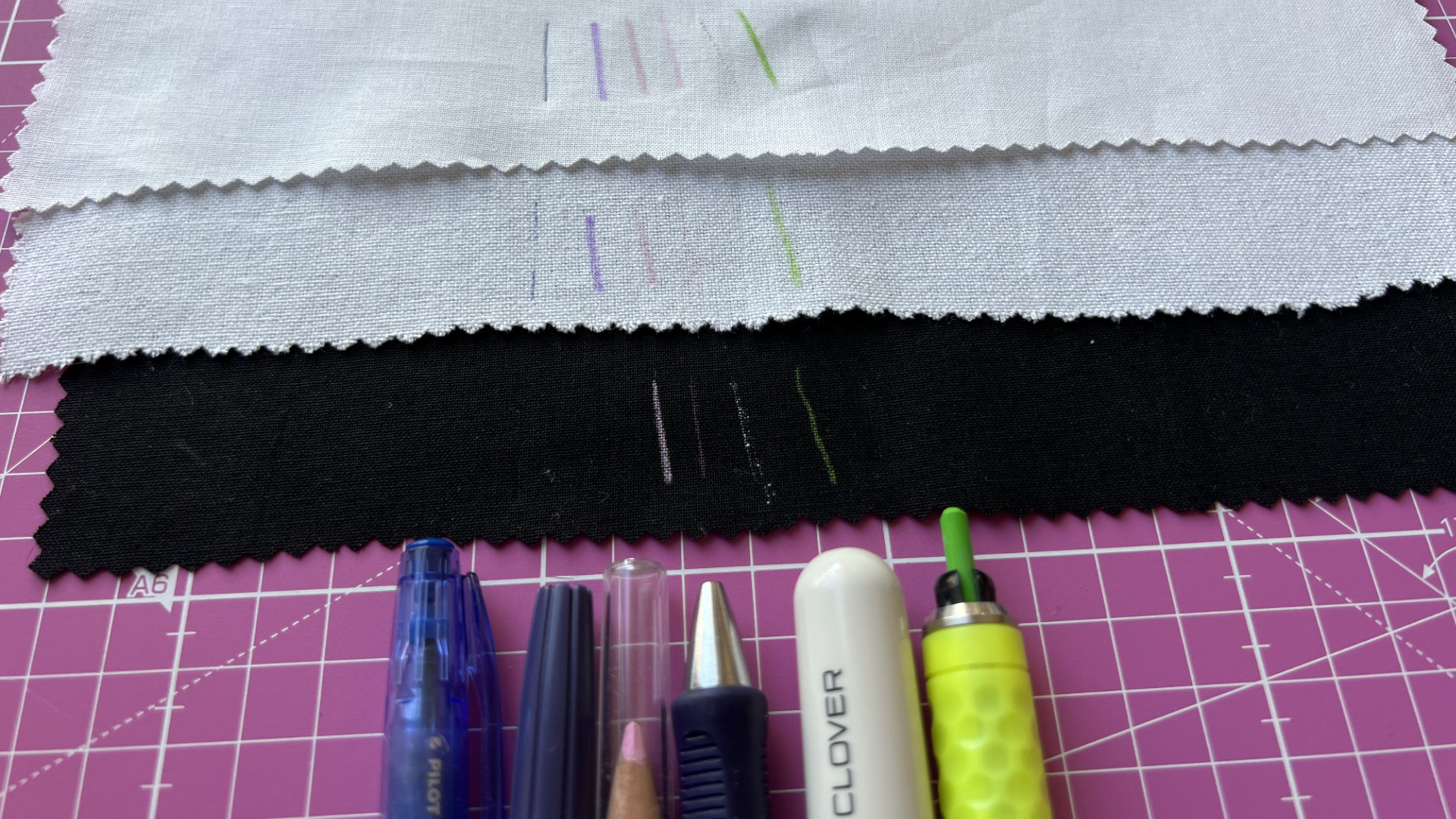 Three zigzag-edged fabric swatches (white and black) on a pink cutting mat with embroidery marks; marking tools in front.