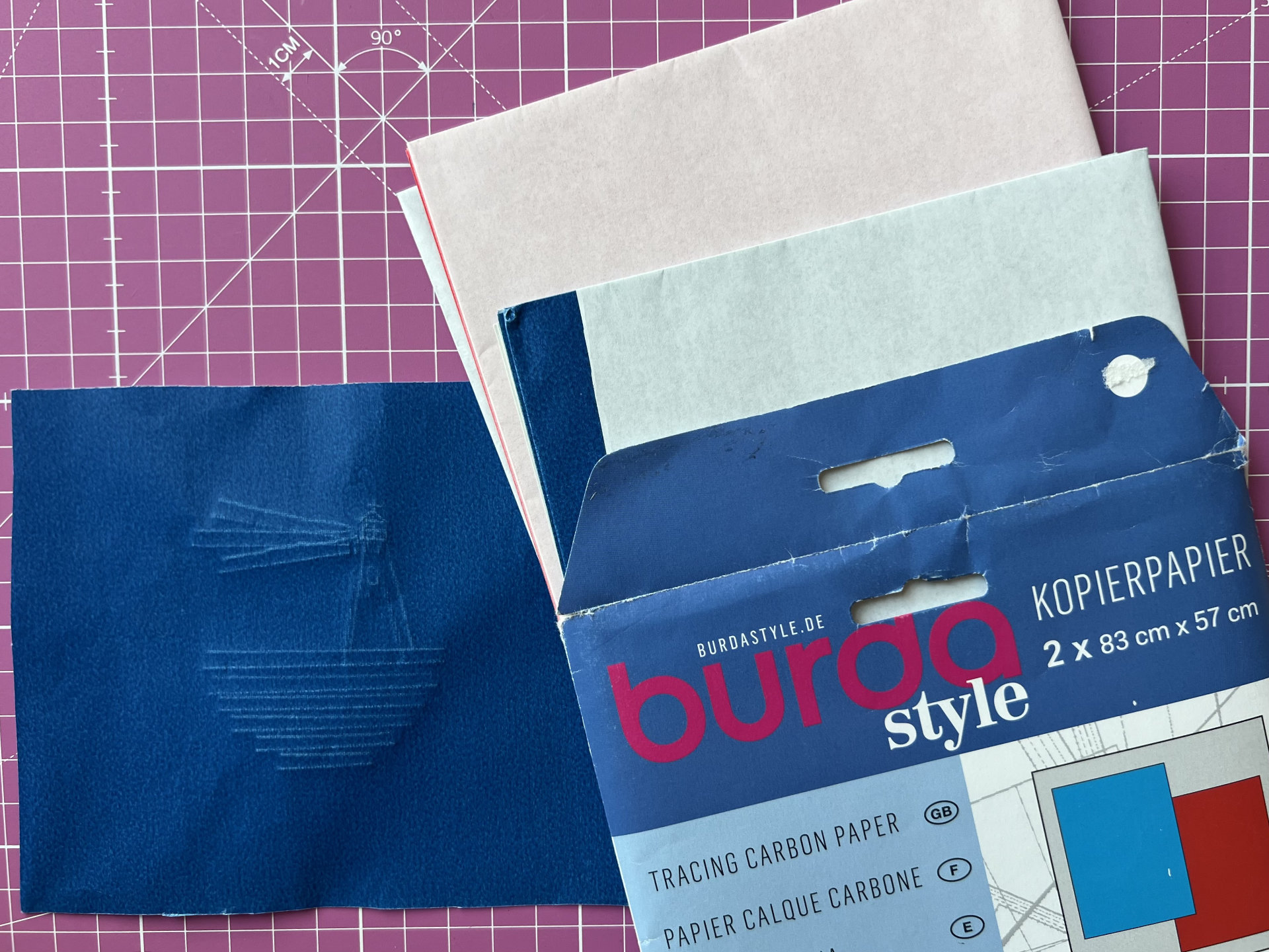Blue tracing carbon paper swatch beside Burda Style packaging on a pink cutting mat.