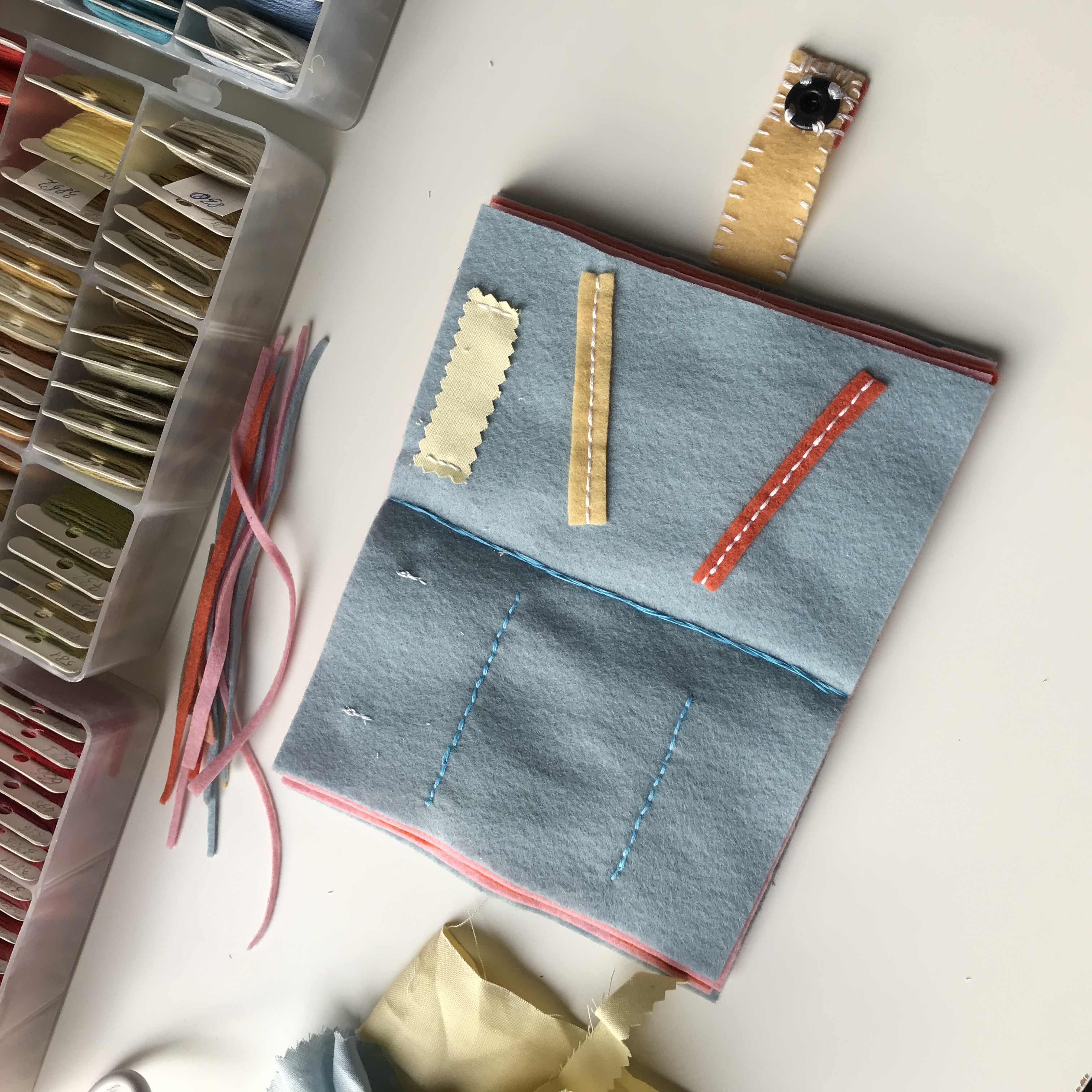 Open blue felt notebook with stitched pockets and ribbons; sewing supplies visible in background.