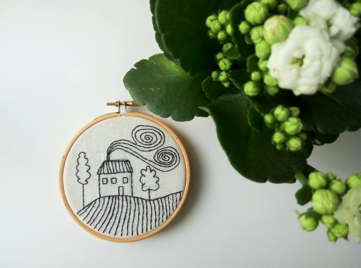 Hand-stitched embroidery in a wooden hoop showing a house on rolling hills with trees and swirly clouds.