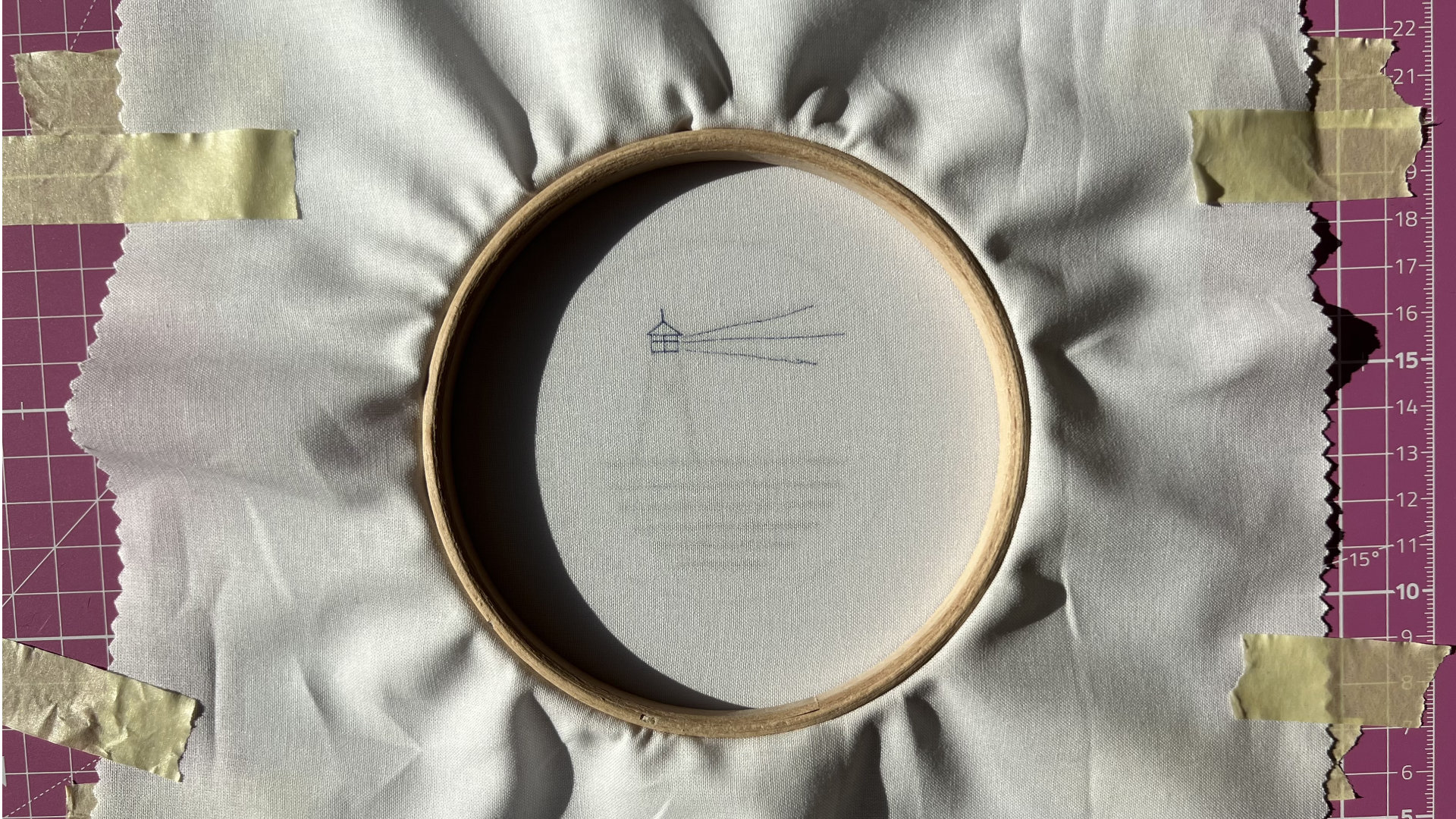 Embroidery hoop on fabric stretched over a pink cutting mat; small blue lighthouse sketch with radiating lines.