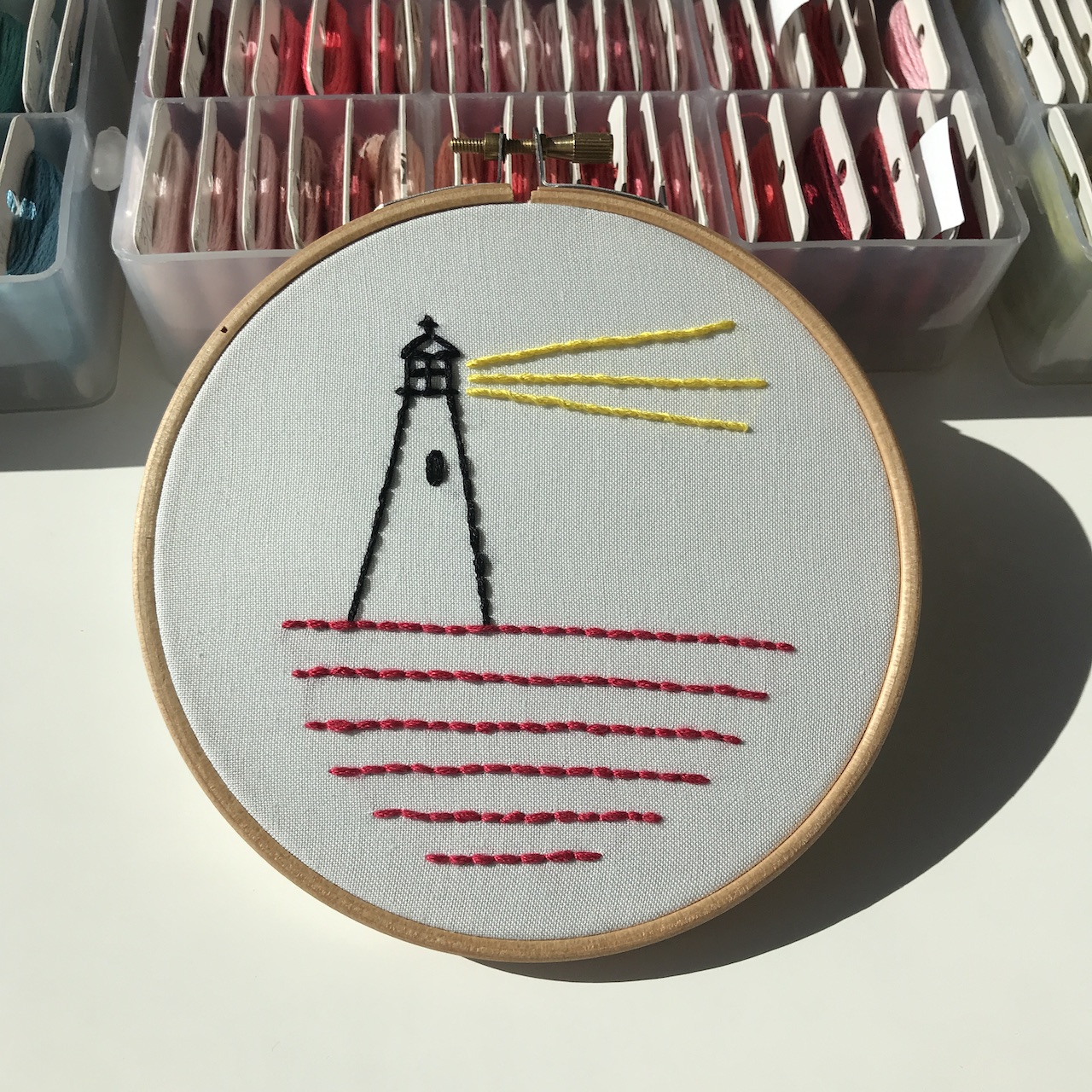 Embroidery hoop with a simple lighthouse: black tower, yellow sun rays, and red waves.