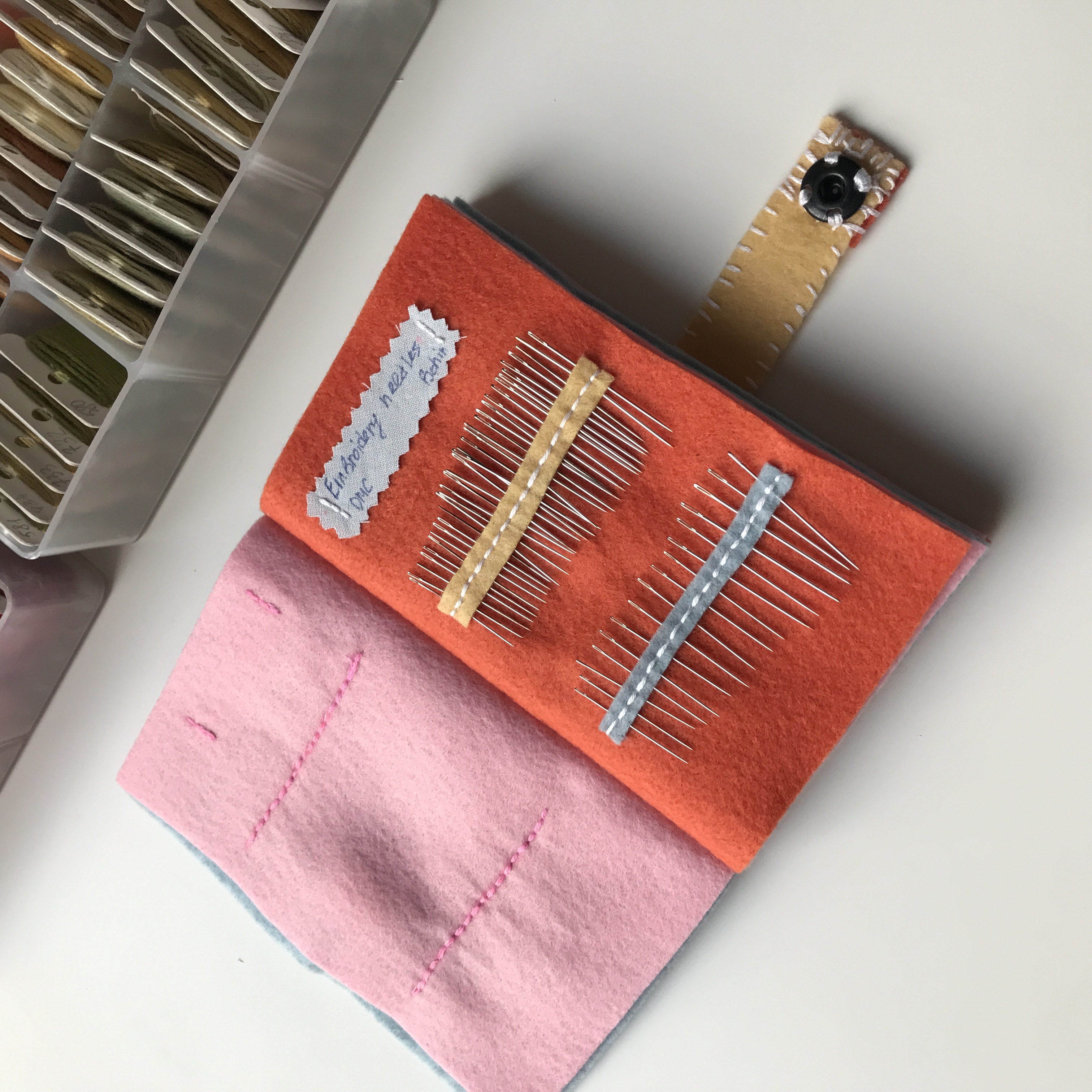 Open embroidery needle book with rows of needles on orange felt, pink pocket page beside it.