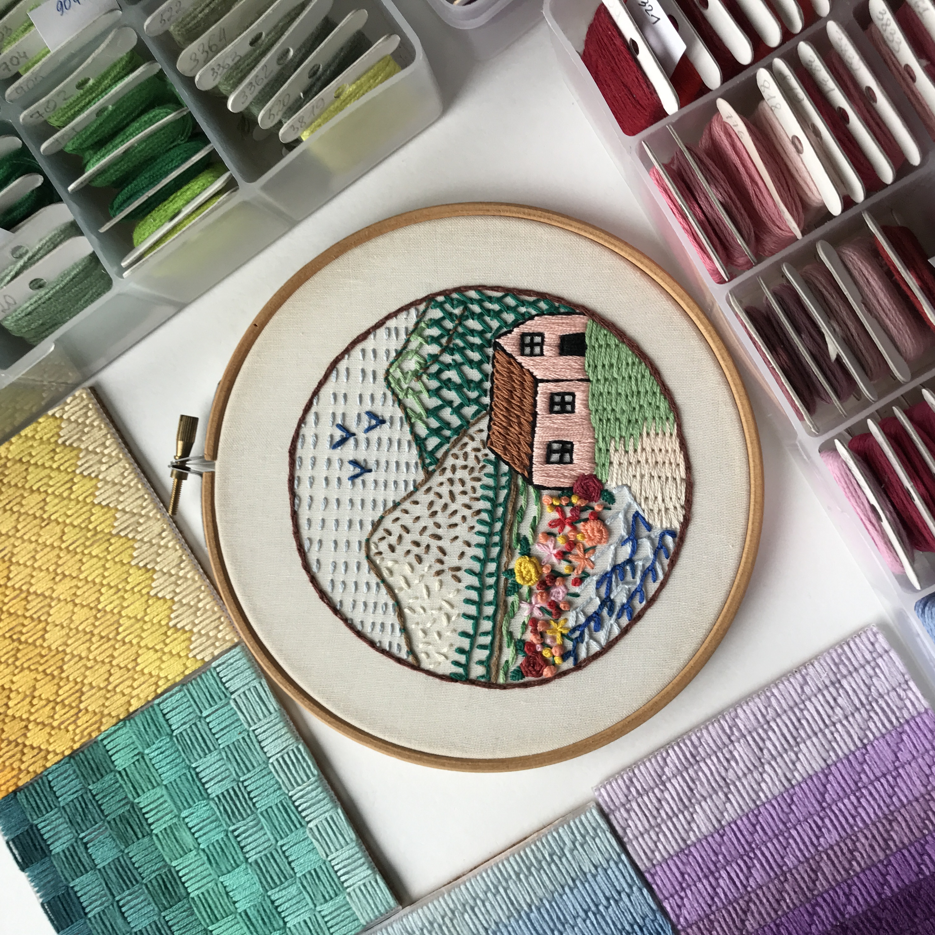 Circular embroidery landscape with mountains, a pink cottage, colorful flowers, and birds.