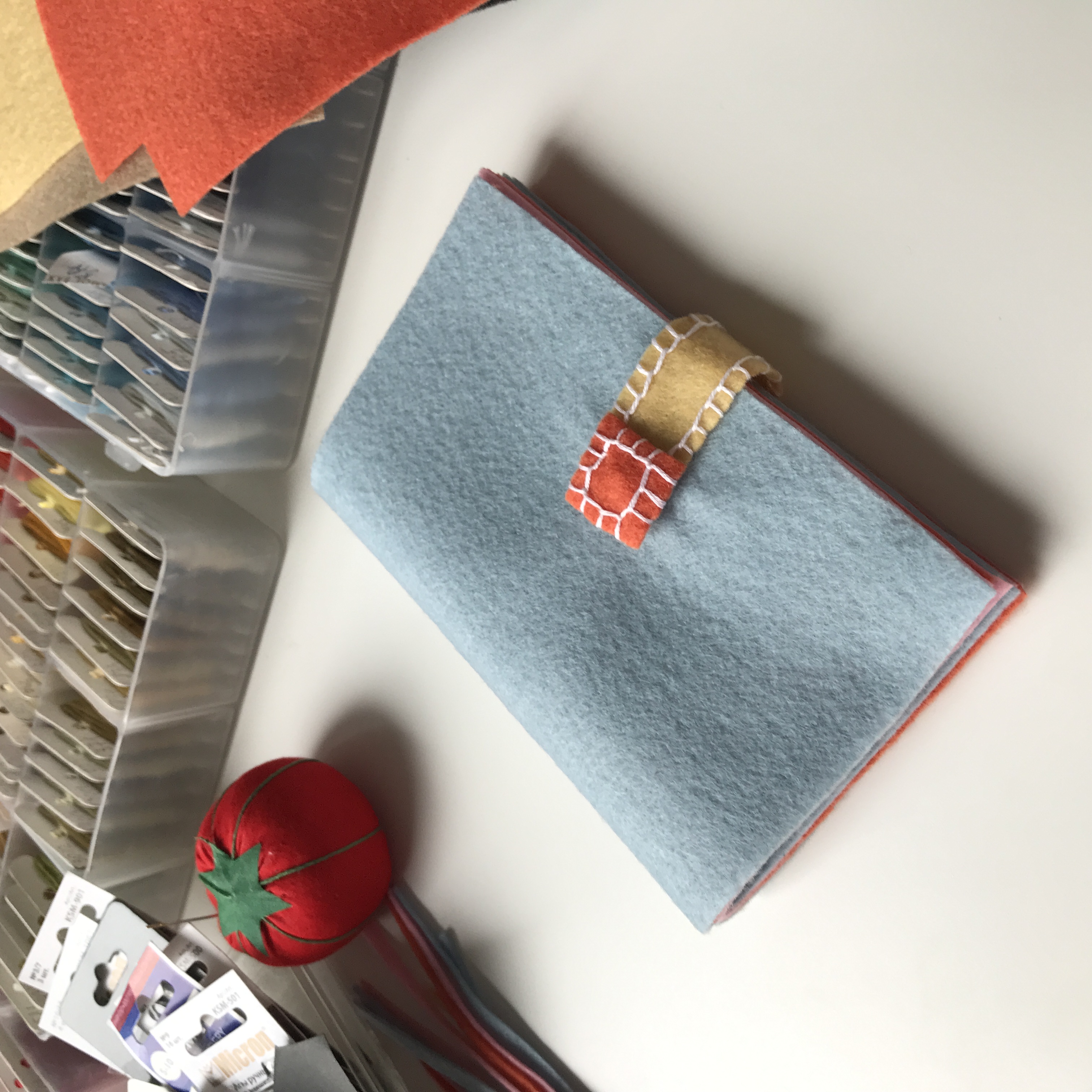Blue felt-covered notebook with a decorative stitched strap (orange tab and beige band) on a white desk