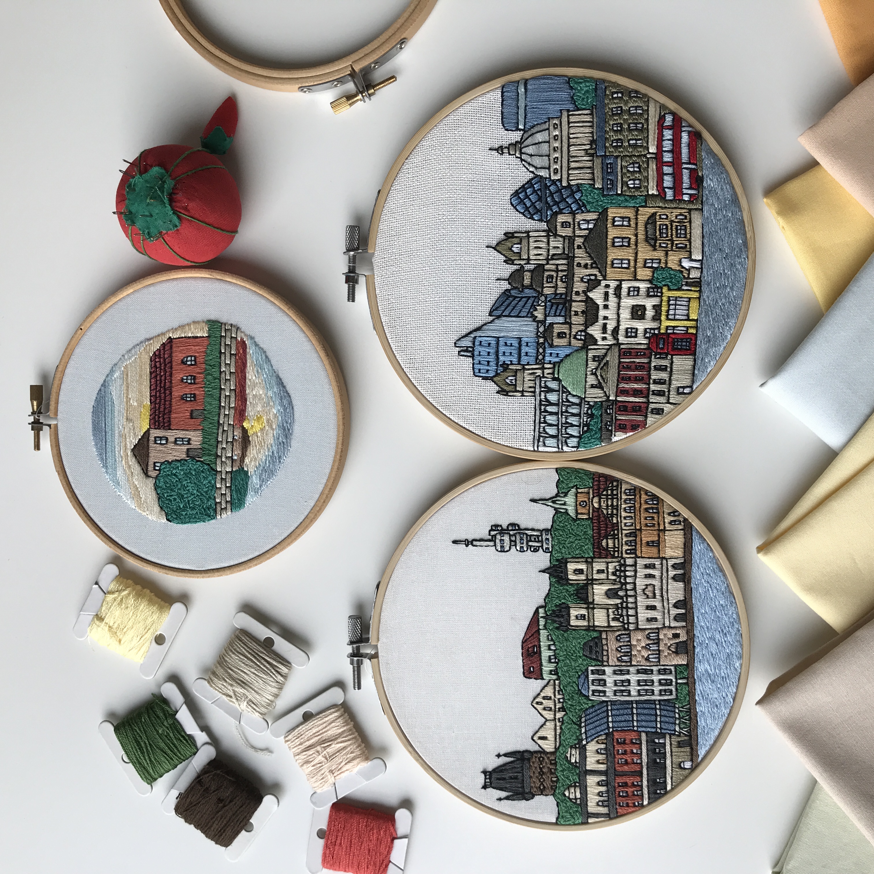 Three embroidery hoops showcasing scenic town and city skyline designs, with colorful thread bobbins nearby. Embroideries from the course Draw and embroider your city by @faimyxstitch