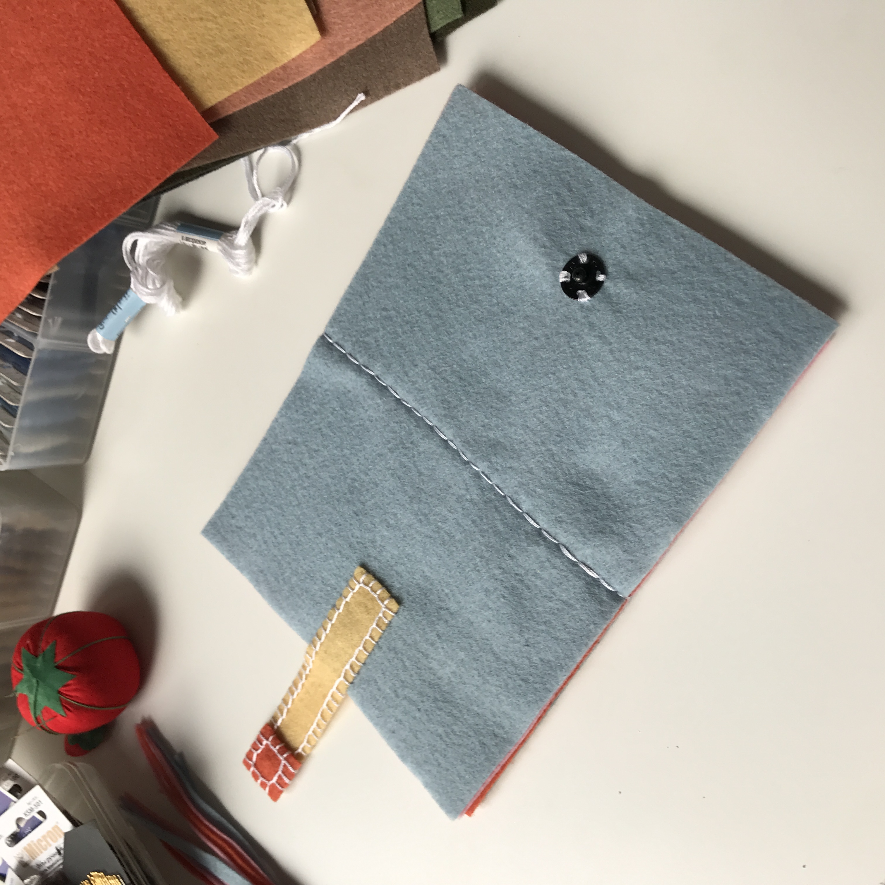 Blue felt needle book with a vertical white back stitch seam, beige tab, and a small black snap button.