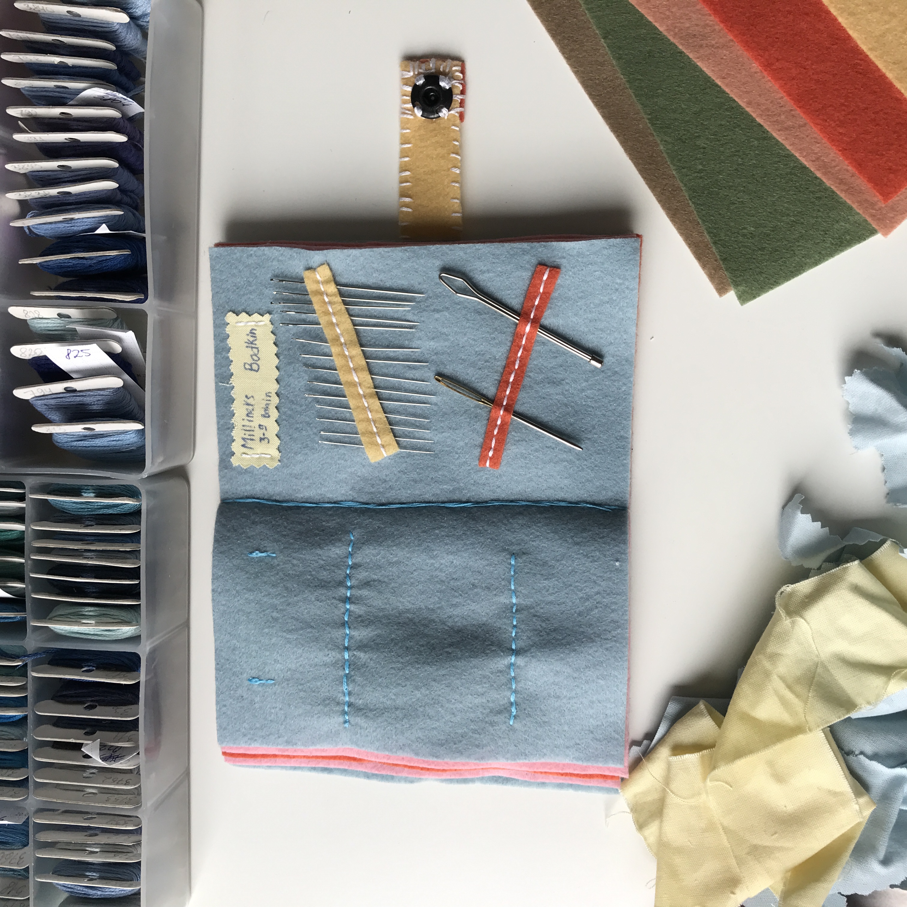 Blue felt mini sewing book opened to show needles, pins, and stitched pockets on a craft desk.